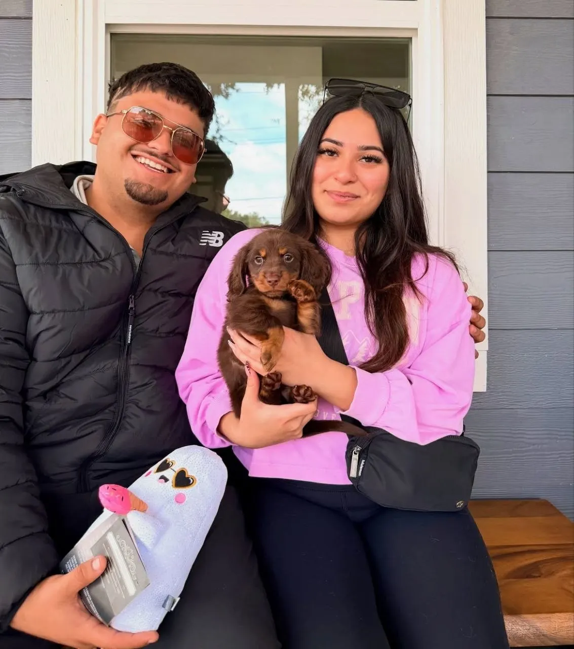 Couple sitting on a front porch step holding a chocolate and tan long-haired Miniature Dachshund puppy with a new toy