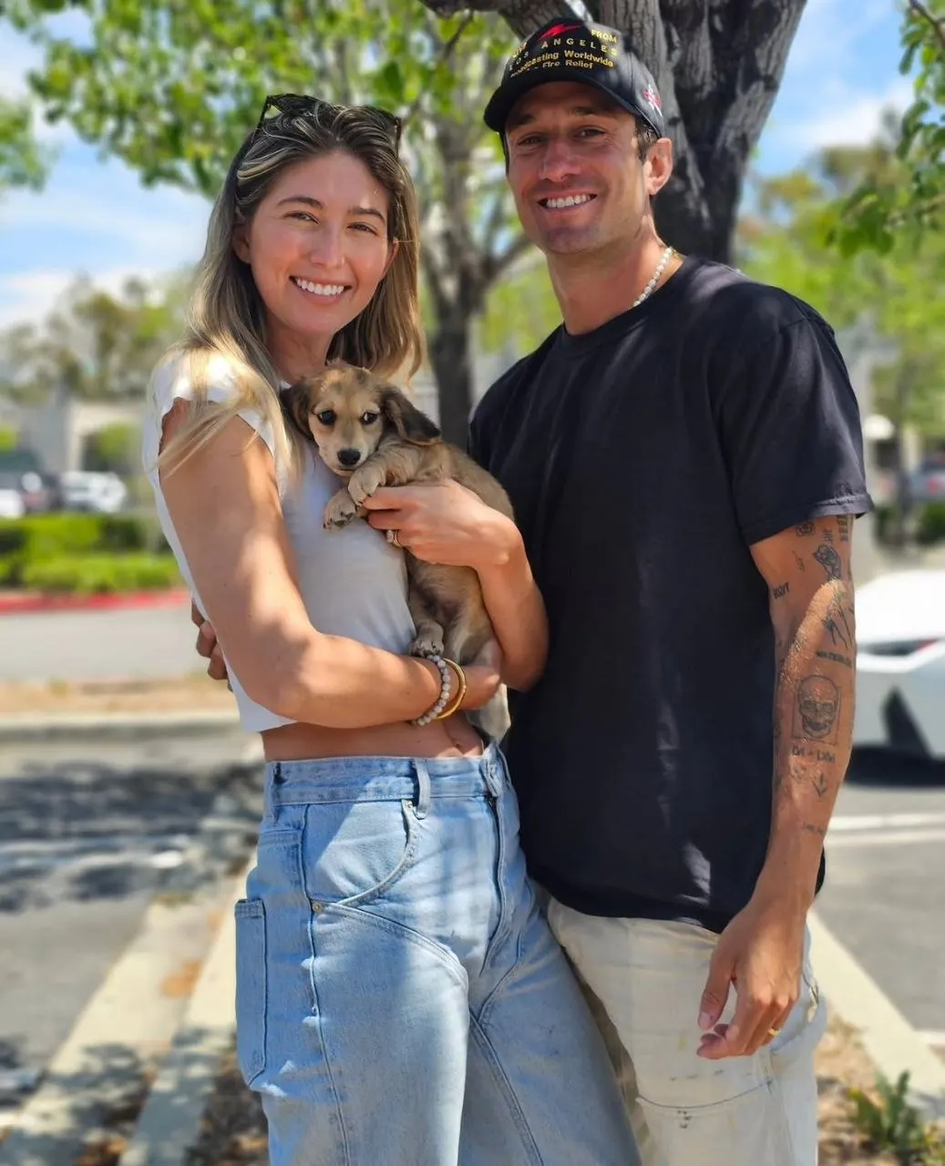 Two people smiling outdoors while holding a shaded cream Miniature Dachshund puppy wrapped in their arms