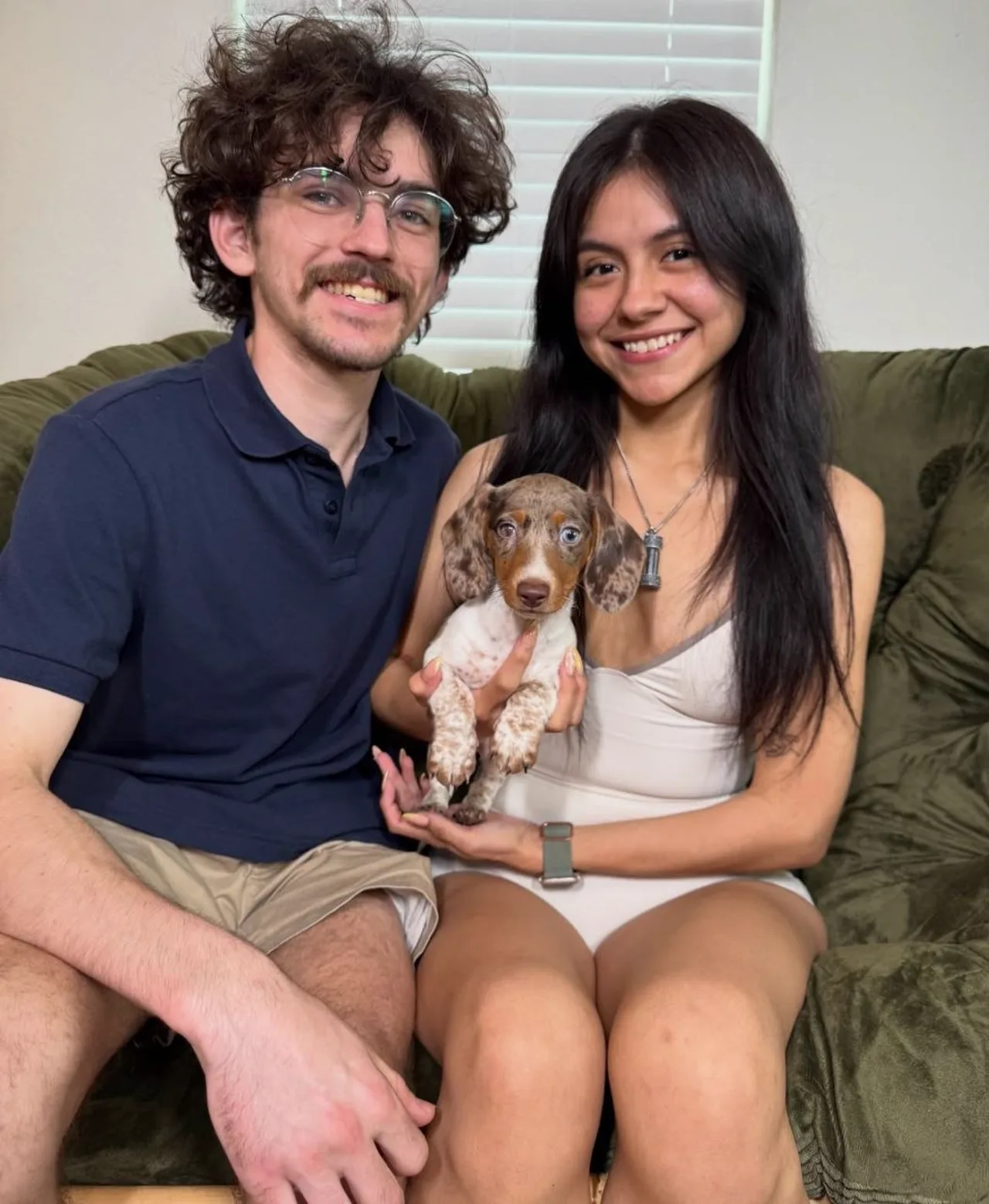Smiling couple seated on a couch holding a chocolate piebald Miniature Dachshund puppy