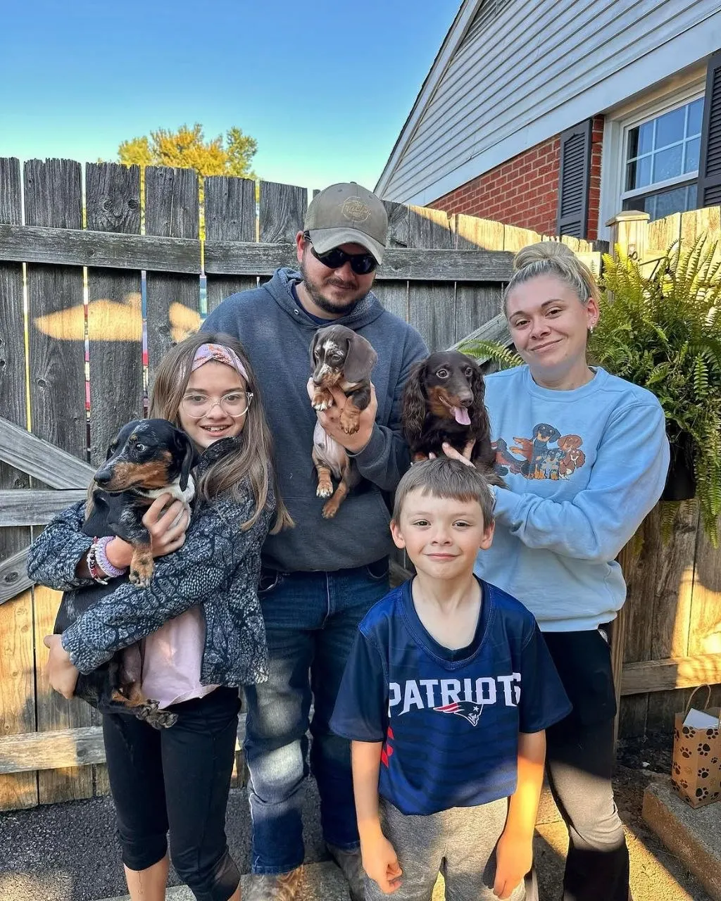 Family of four standing in a backyard holding three Miniature Dachshunds, including a small dapple puppy