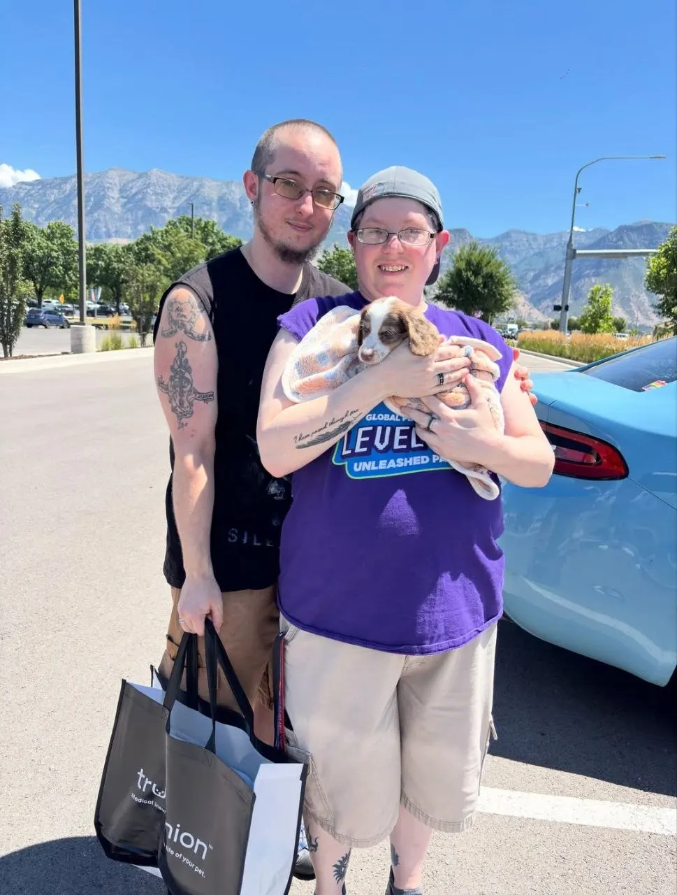 Two people holding a piebald Miniature Dachshund puppy wrapped in a blanket, with mountains behind them